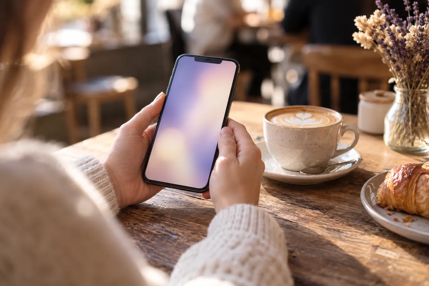Hands holding a phone at a café table beside a cappuccino and a croissant.